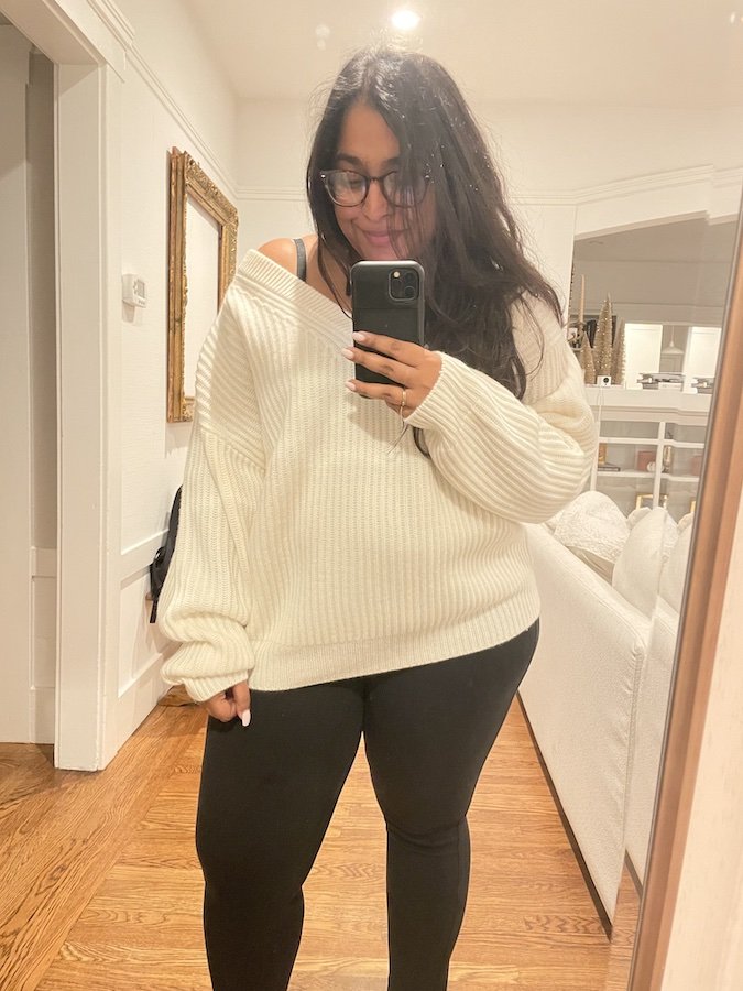 Woman with long dark hair wearing glasses, a white oversized sweater, and black leggings takes a mirror selfie in a well-lit room with wooden floors and white walls.