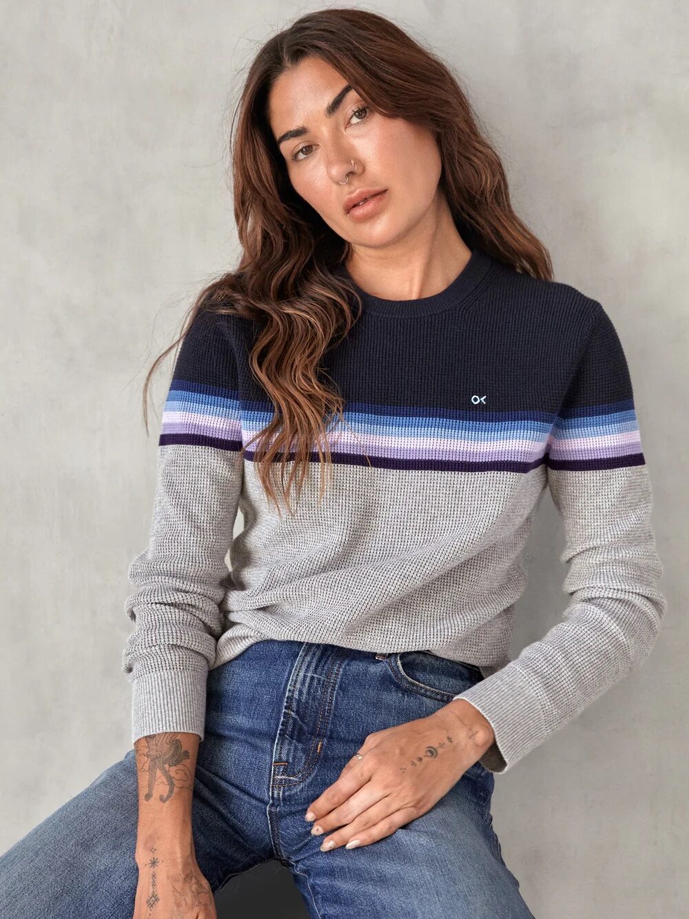 Woman with long brown hair wearing a gray sweater with navy, blue, and purple stripes, and blue jeans, sitting against a plain light-colored background.