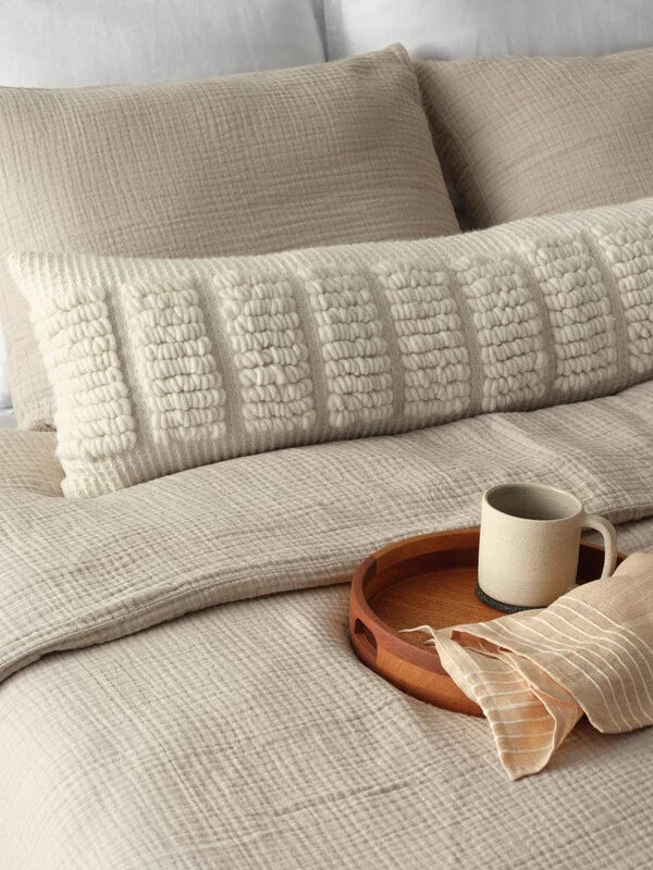 A neatly made bed with textured beige bedding, a decorative long pillow, and a wooden tray holding a beige mug and folded napkin.