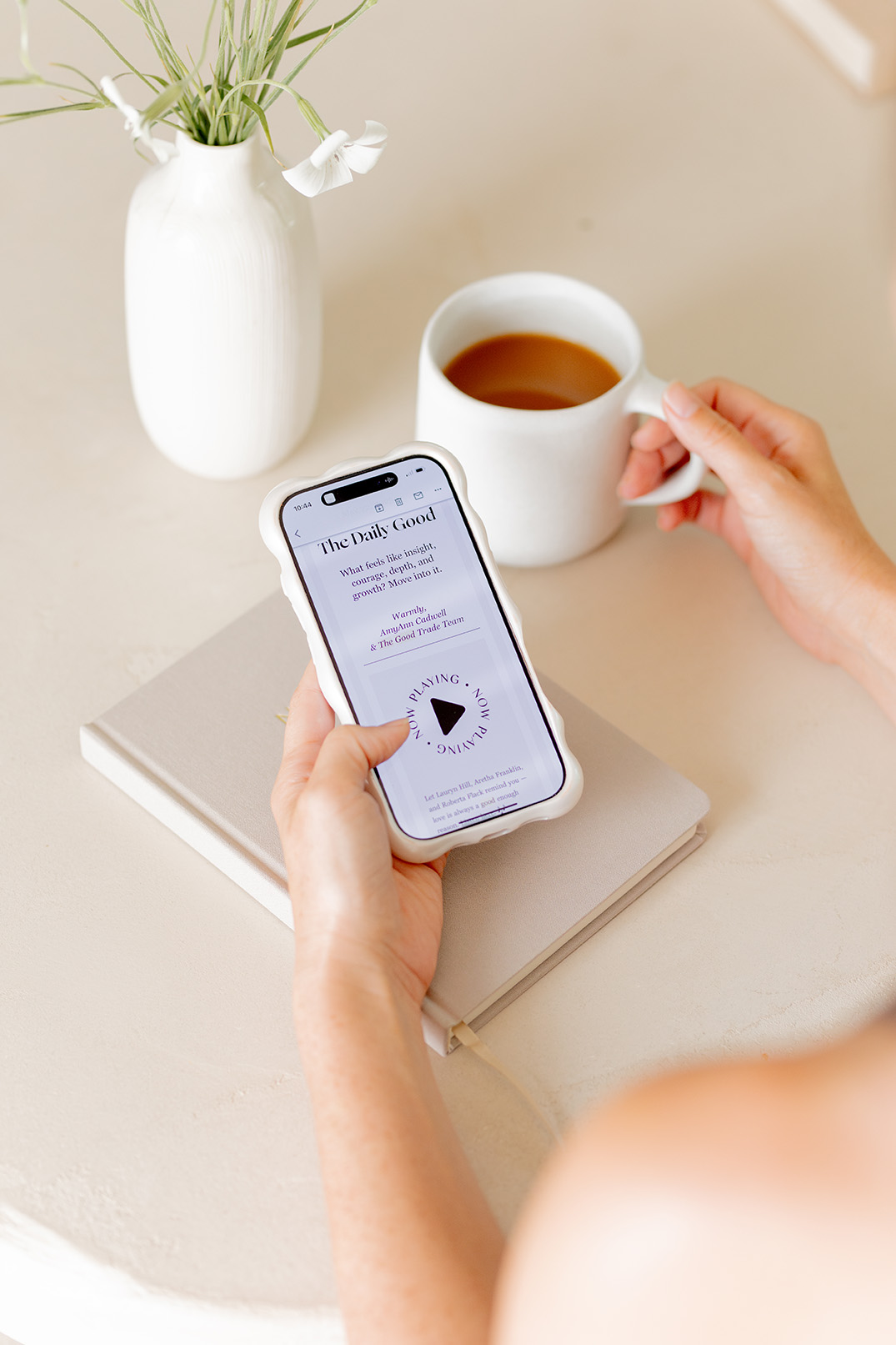A person holding a smartphone displaying an article while sitting at a table with a cup of tea or coffee, a closed notebook, and a white vase with green stems.