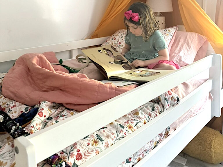 A young girl sits on a bed with floral bedding and an organic mattress, reading a book under a yellow canopy decorated with colorful tissue paper pom-poms.