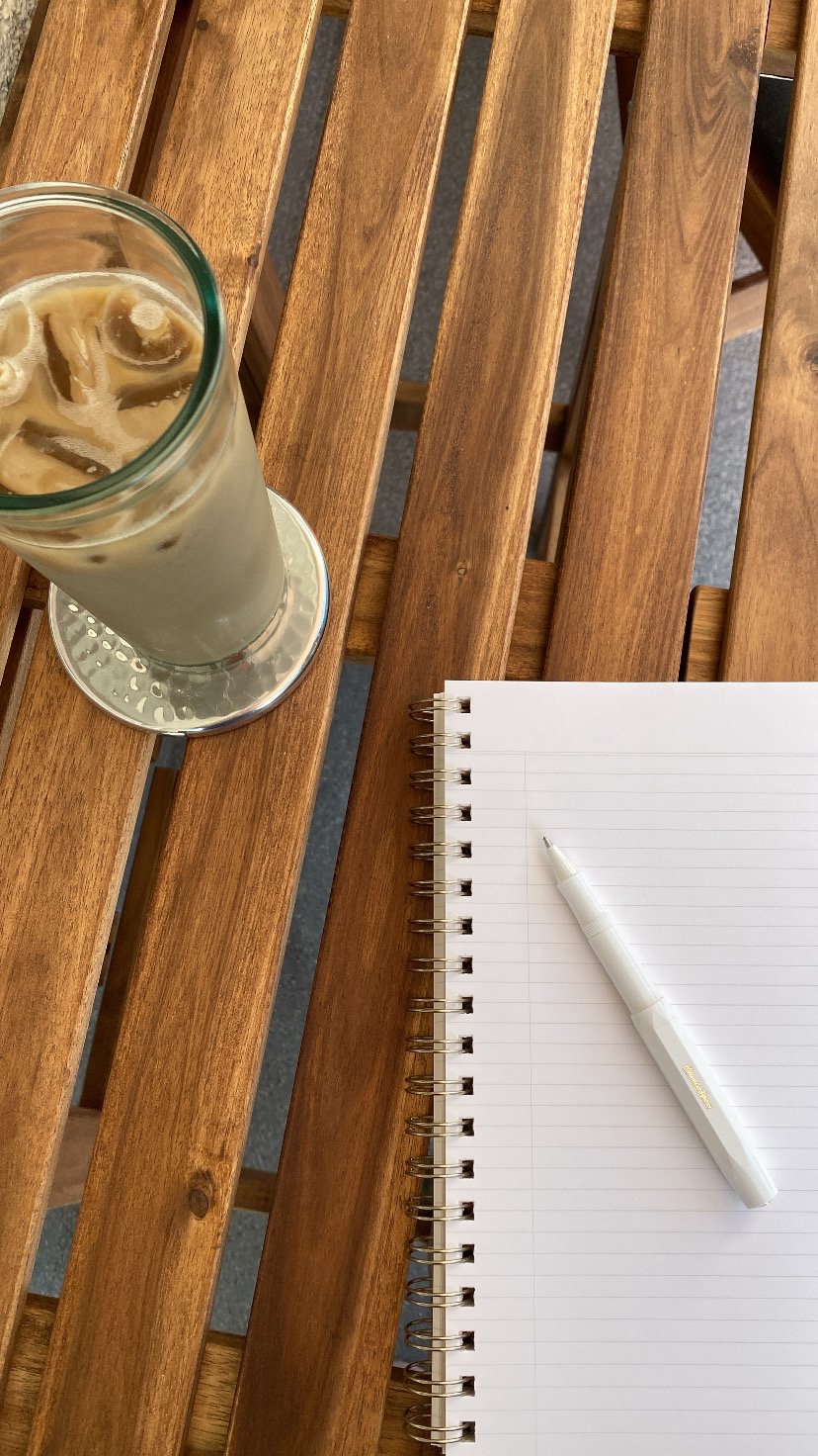 A glass of iced coffee, a spiral notebook with blank lined paper, and a white pen are on a wooden table.