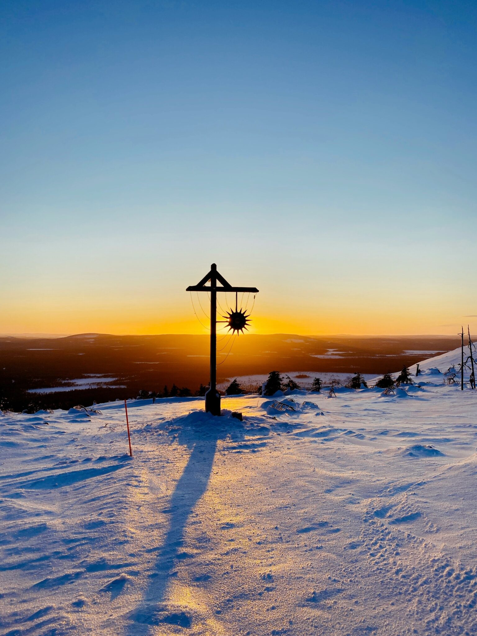 A wooden post with a metal sun ornament casts a long shadow on a snowy landscape at sunset, with distant trees and a clear sky in the background.