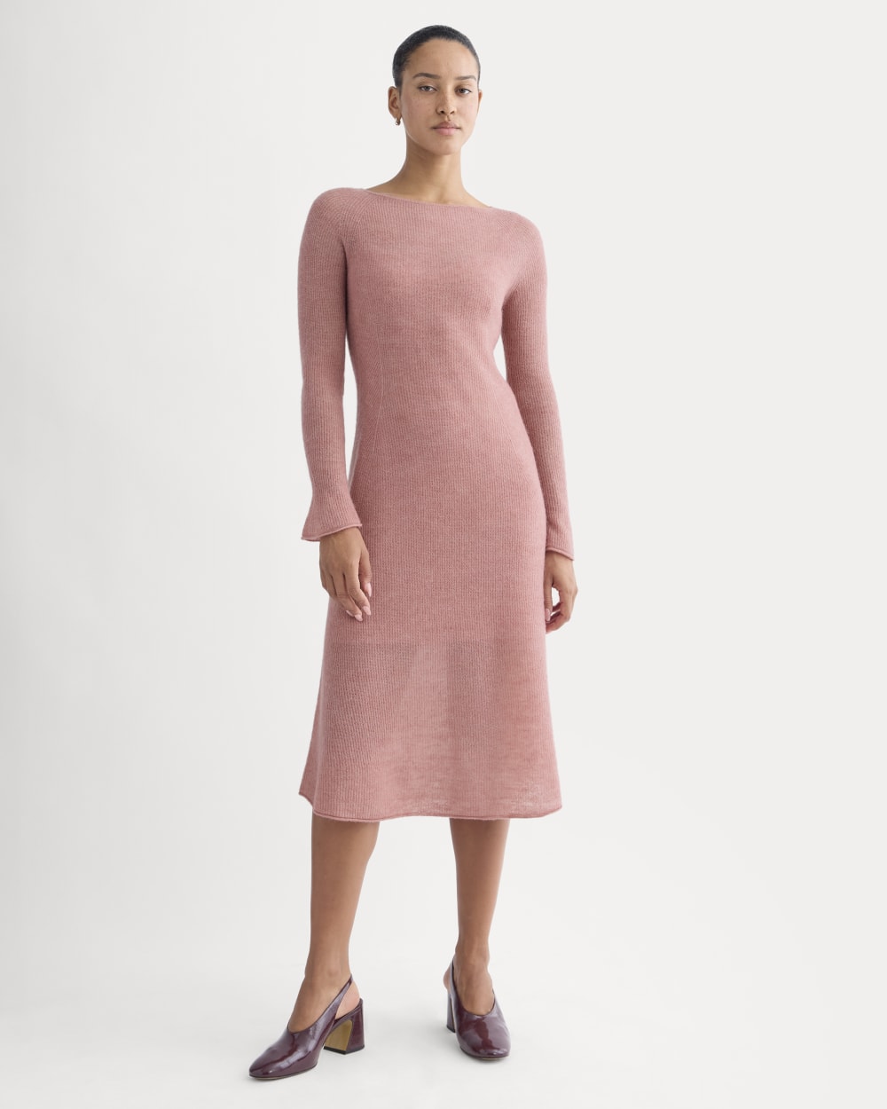 A woman stands against a plain background wearing a long-sleeve, light pink knit midi dress and burgundy block-heeled shoes.