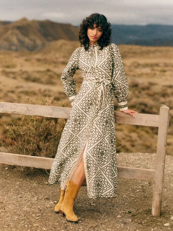 Woman wearing a long patterned dress and tan boots stands by a wooden fence in a dry, mountainous landscape.