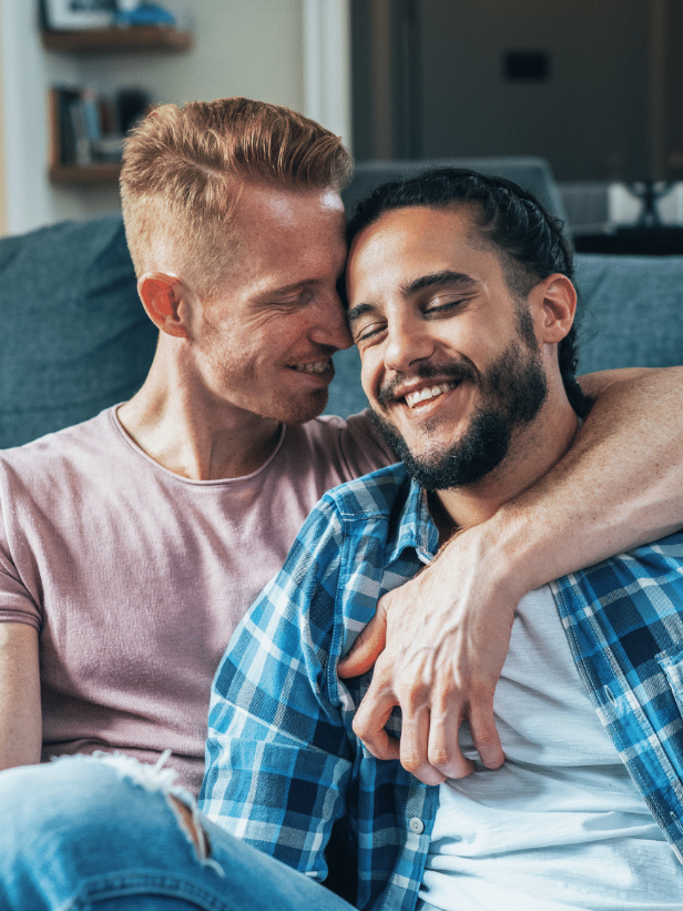 Two men sitting on a couch, one in a pink shirt and the other in a blue plaid shirt, smiling and embracing affectionately.