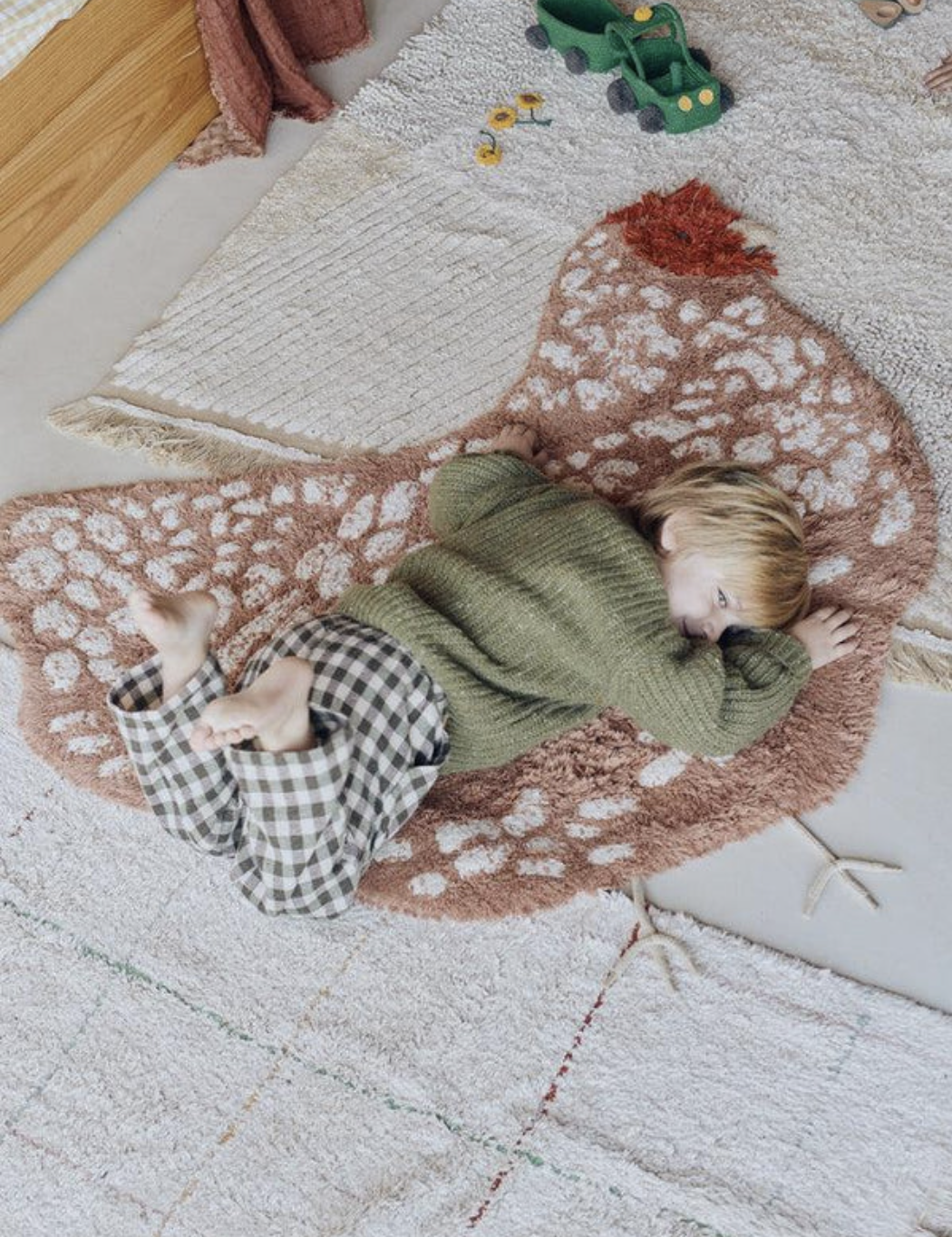 Child lying on a chicken-shaped rug, wearing a green sweater and plaid pants, surrounded by toys on a carpeted floor.