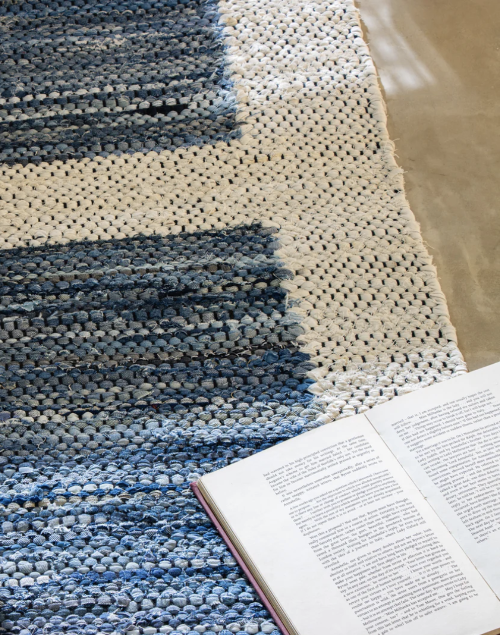 Open book on a polished floor next to a textured, striped rug in shades of blue and white.