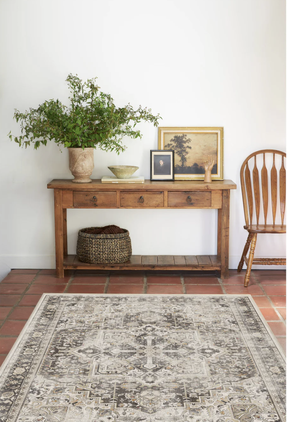 Wooden console table with plants, framed pictures, and decorative items. A wicker basket is underneath. A patterned rug lies on terracotta tiles next to a wooden chair.