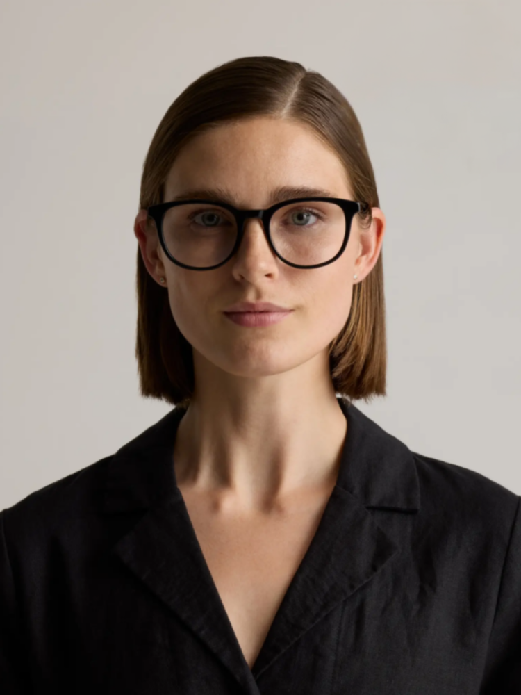 A woman with straight brown hair, wearing black-rimmed glasses and a black blazer, stands against a plain light background, looking directly at the camera.