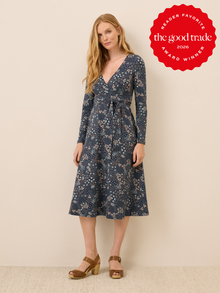 Woman wearing a long-sleeve, floral-patterned, blue wrap dress with a tie waist and tan platform sandals, standing against a beige background. "The Good Trade Award Winner 2026" badge in corner.