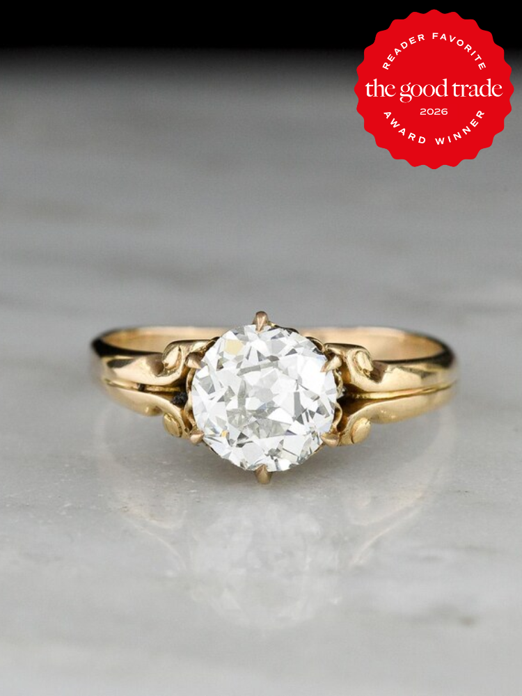 A gold ring with a round, clear gemstone displayed on a marble surface; a red badge in the corner reads “The Good Trade Reader Favorite Award Winner 2026.”.