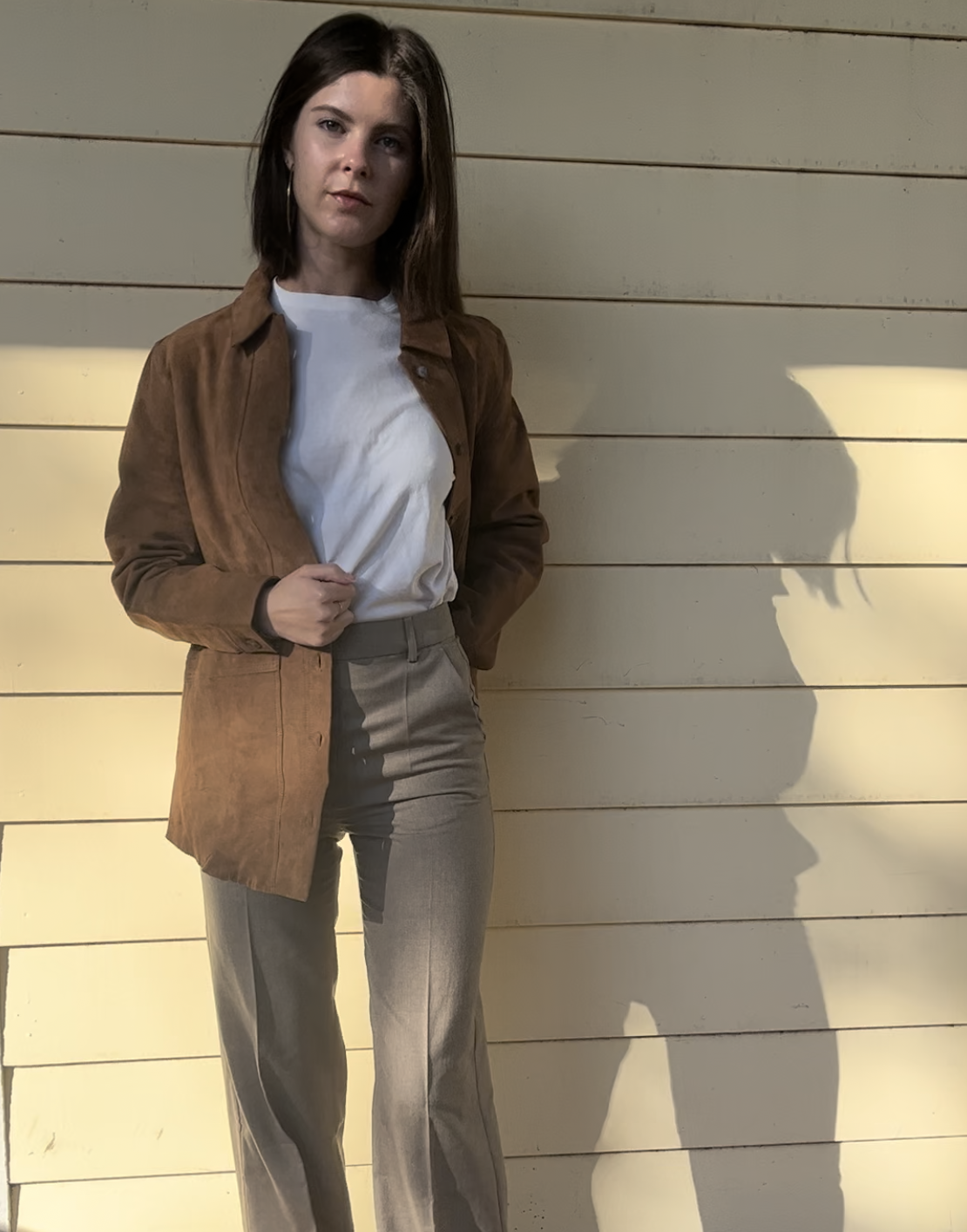 A person stands on a wooden deck against a cream paneled wall, wearing a brown jacket, white t-shirt, beige pants, and black shoes in sunlight.