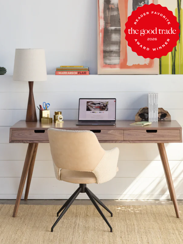 A modern wooden desk with a lamp, books, and office supplies, a beige swivel chair, and a laptop, set against a white shiplap wall with abstract art and a red award badge in the corner.