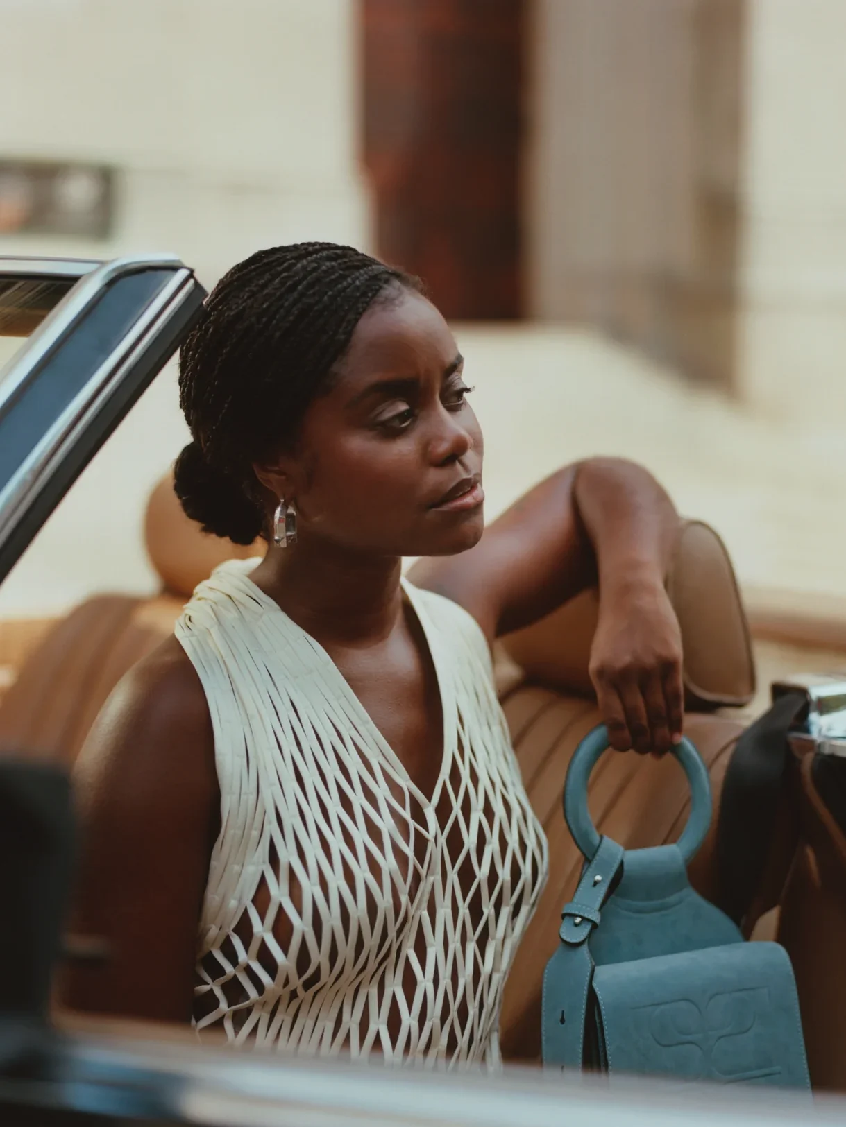 Person sitting in a convertible car wearing a white netted top, holding a blue handbag, looking thoughtfully into the distance.