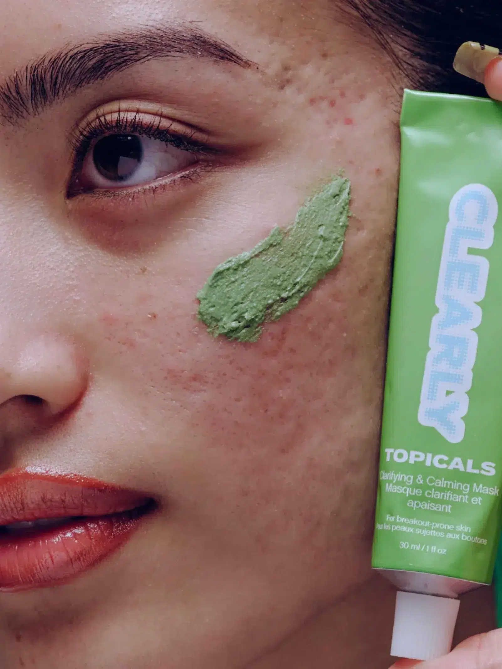 Close-up of a person applying a green clarifying and calming mask on their cheek, with a tube labeled "Topicals" beside their face.