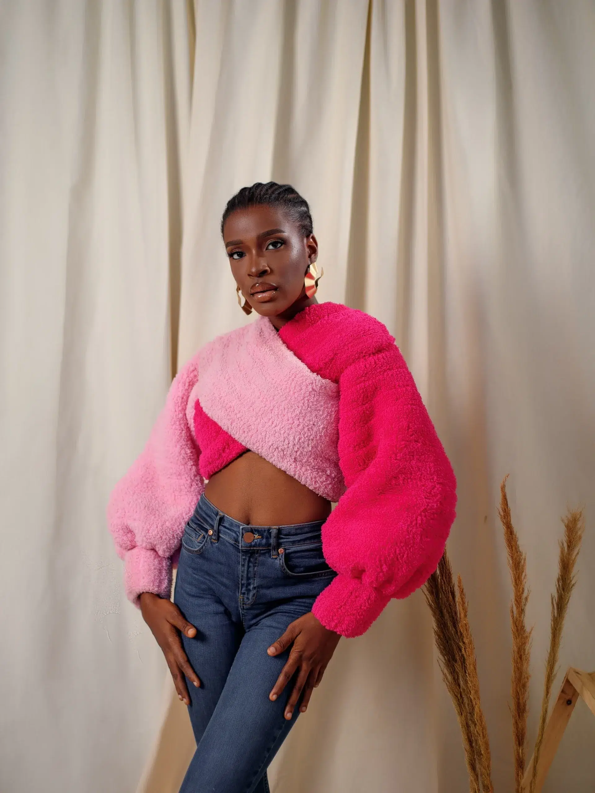 Person wearing a pink and red textured sweater, jeans, and large earrings, stands in front of a curtain with dried plants beside them.