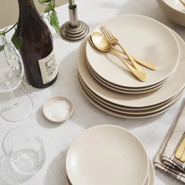 A stack of beige ceramic plates with gold cutlery rests on a white tablecloth beside a wine bottle, a glass, a candle holder, a bowl of salt, and folded napkins.