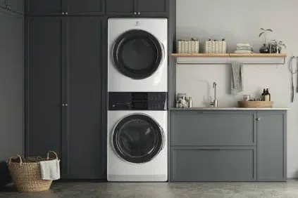 A modern laundry room with a stacked washer and dryer, dark gray cabinets, a wicker laundry basket, and a countertop with a sink and shelves above.