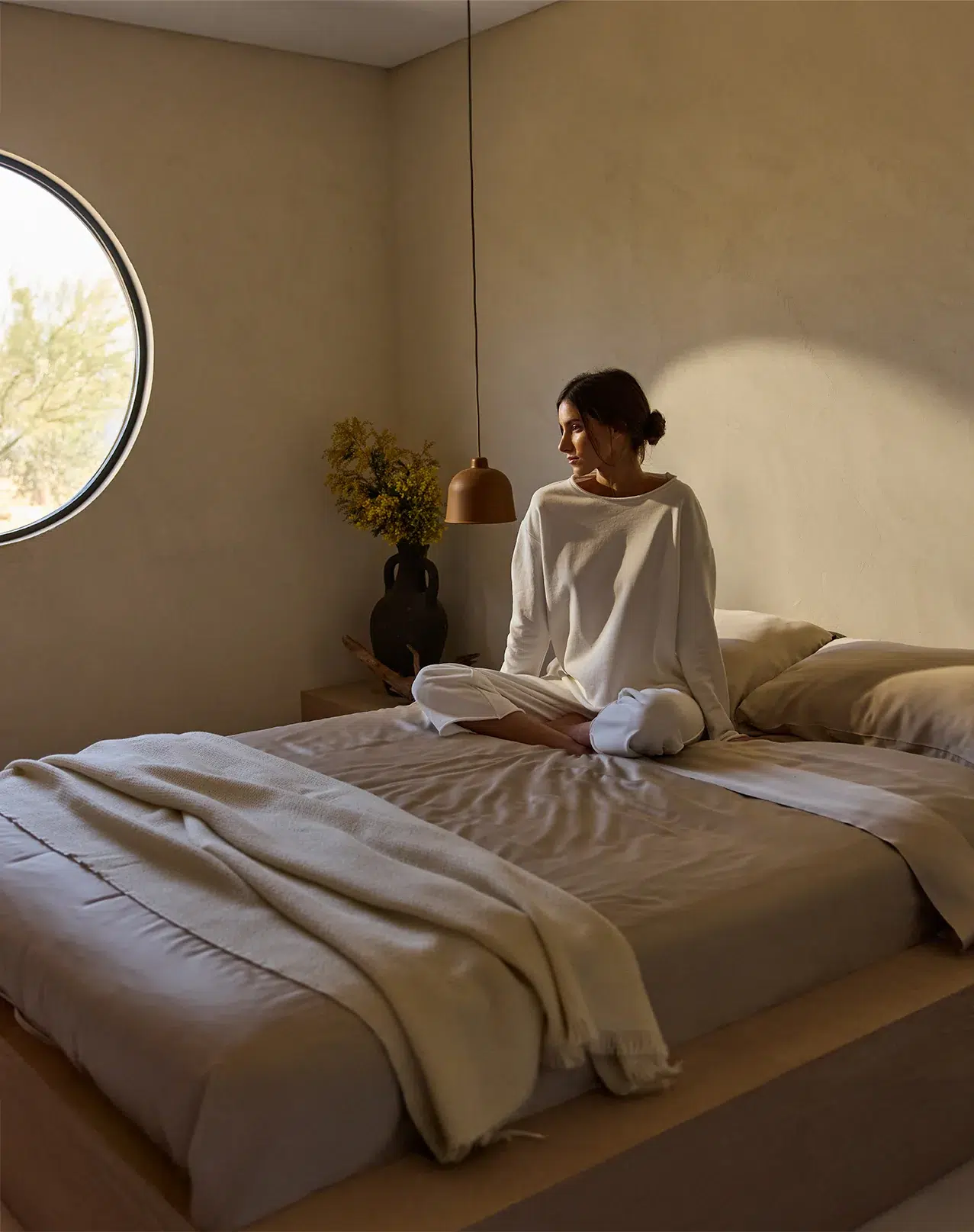A woman in white loungewear sits cross-legged on a neatly made bed in a minimal, sunlit bedroom with beige walls and a round window.