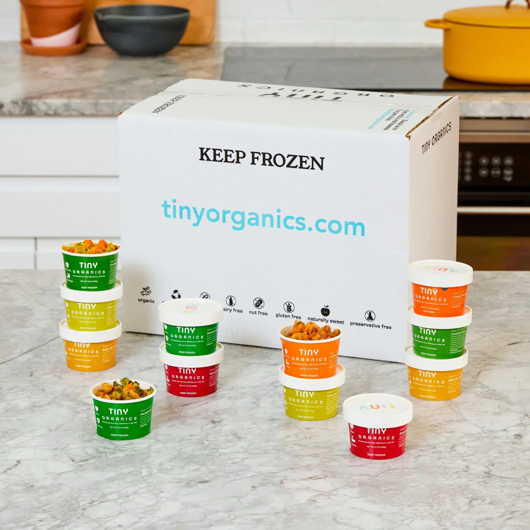 A box labeled "Keep Frozen" from tinyorganics.com on a kitchen counter with 12 assorted Tiny Organics food containers arranged in front of it.