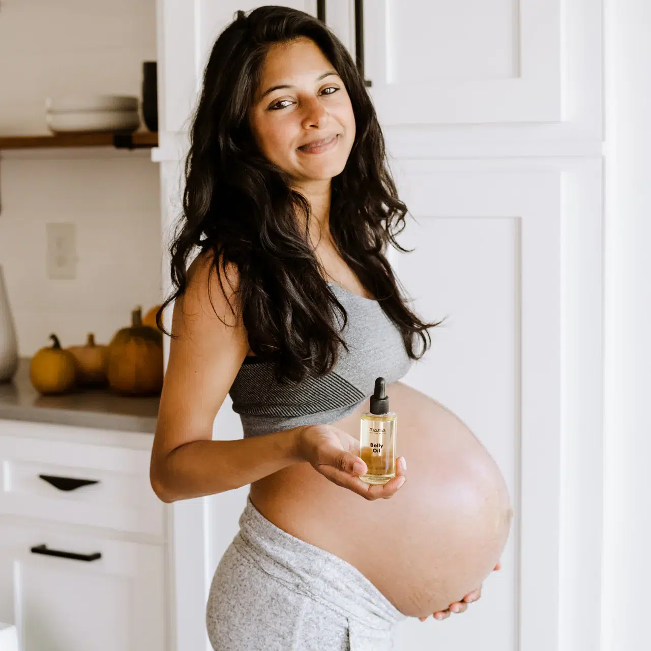 A pregnant woman in athletic wear stands in a kitchen, smiling and holding a small bottle of oil with a dropper.