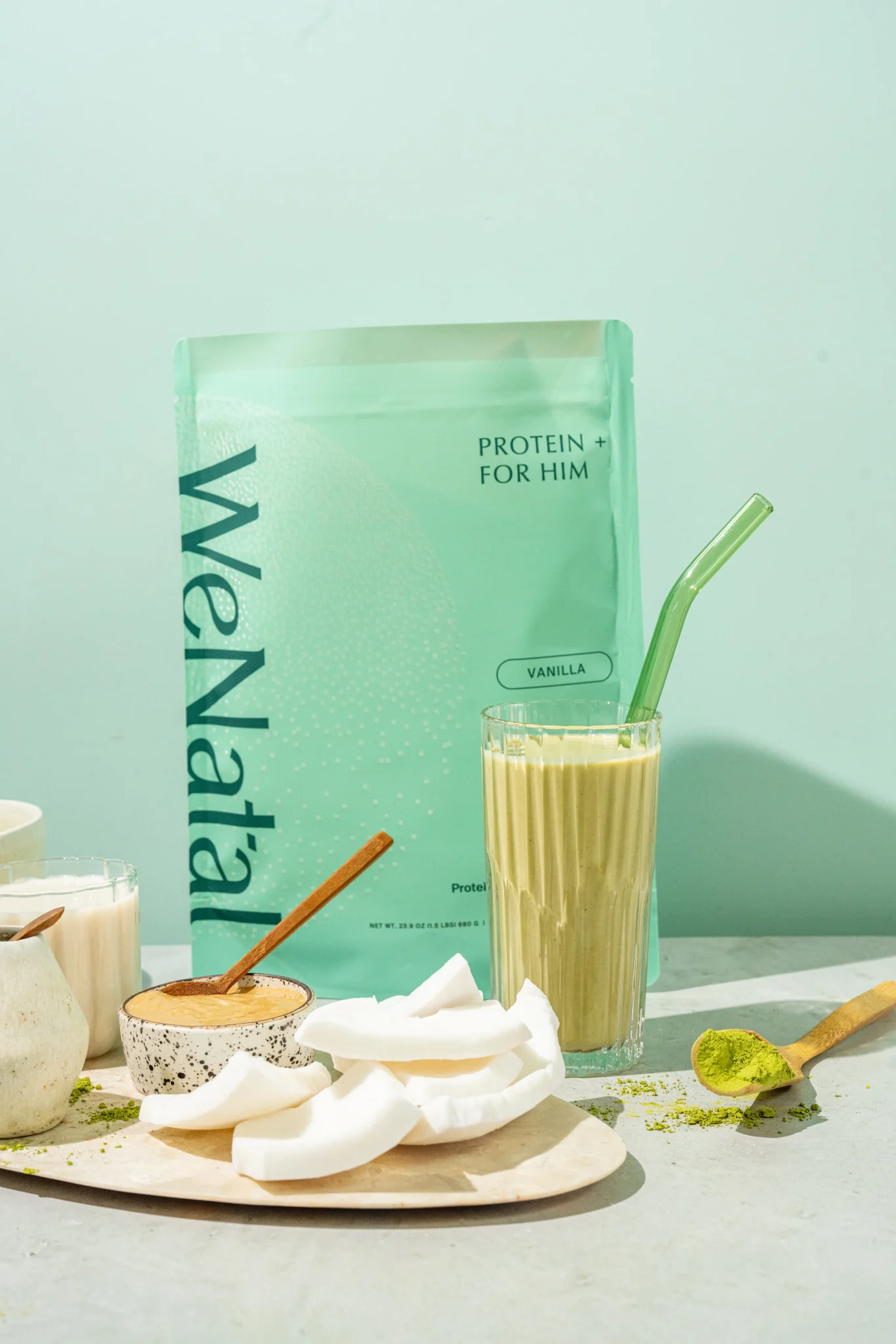 A green protein powder pouch labeled "WeNatal Protein + For Him Vanilla" stands behind a glass of green smoothie, coconut pieces, matcha powder, and other smoothie ingredients.
